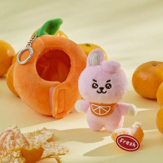 BTS BT21 Cooky Plush toy resembling a pink rabbit character with an orange shaped outfit, surrounded by oranges on a light background.