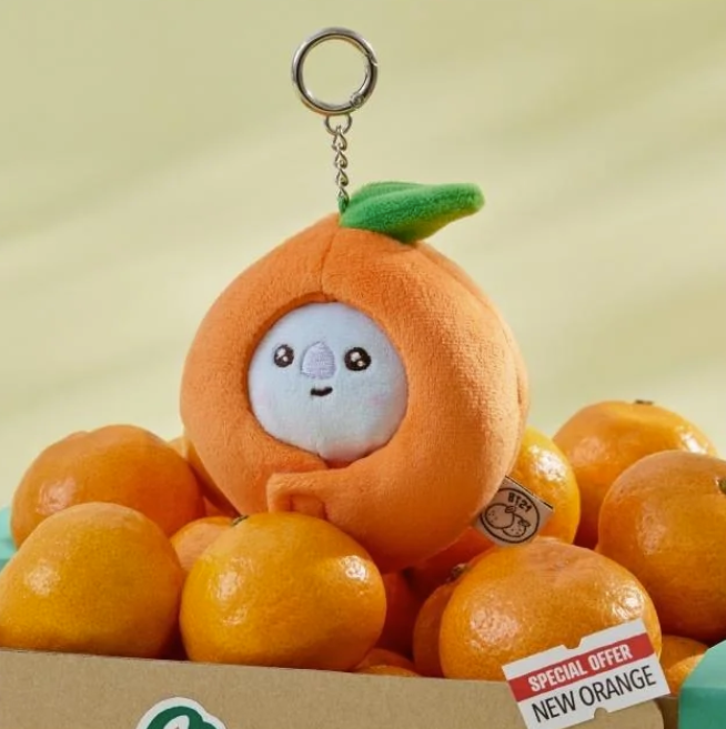 BTS BT21 Koya Plush toy in an orange costume with a keychain, surrounded by real oranges with a 'Special Offer New Orange' sign.