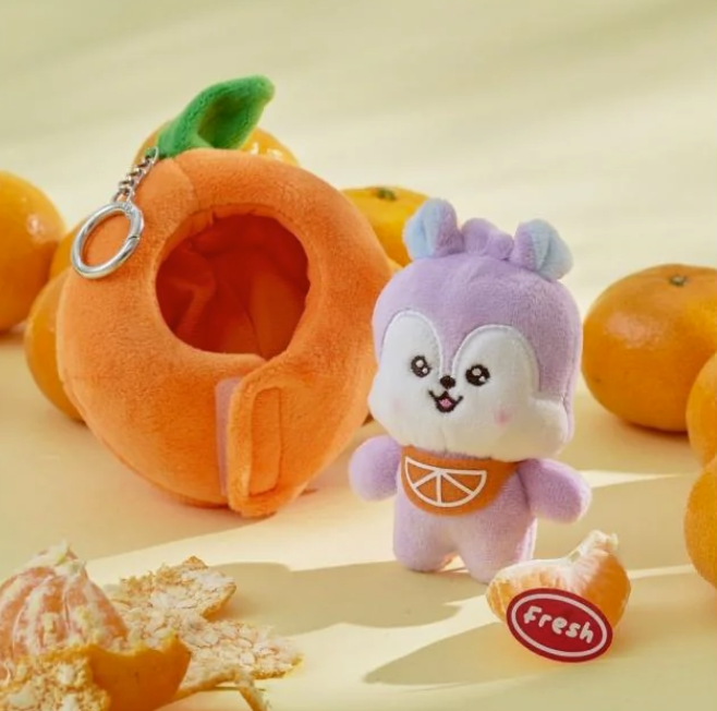 BTS BT21 Mang Plush toy resembling a purple animal with an orange shaped outfit, surrounded by orange fruits on a light background.