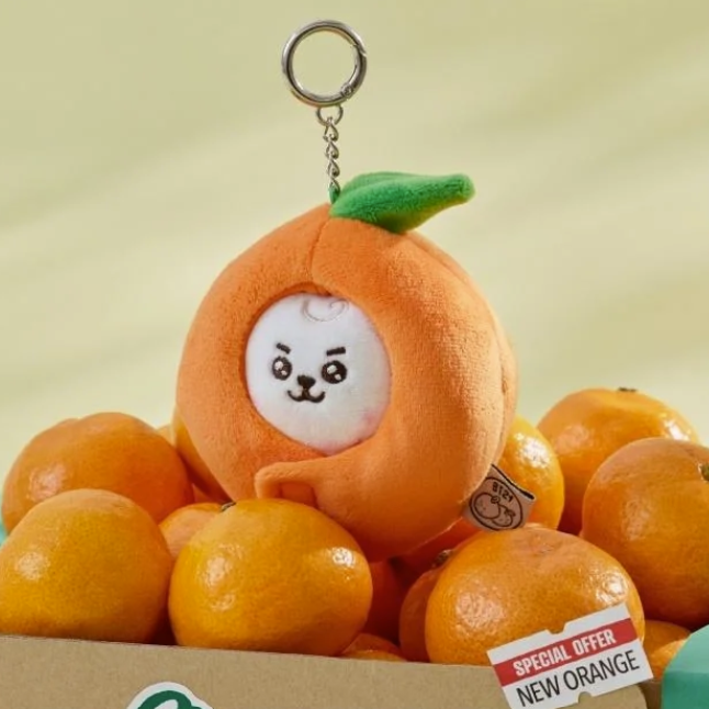 BTS BT21 RJ Plush toy shaped like an orange with a face, surrounded by real oranges, on a light green background.