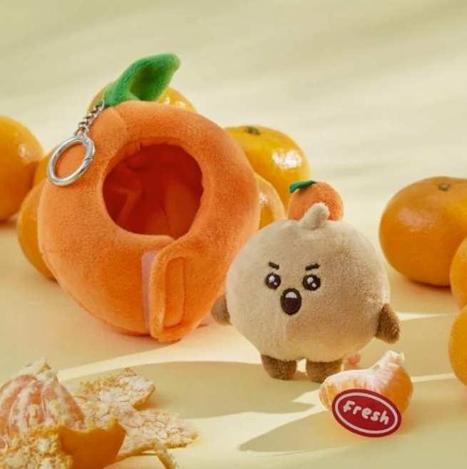 BTS BT21 Shooky Plush toy resembling a small animal with an orange-shaped outfit and mandarin oranges on a beige background