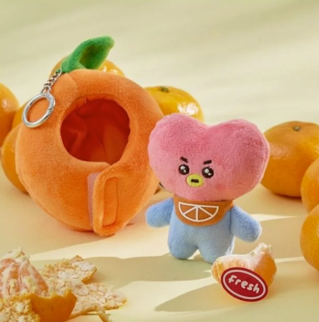 BTS BT21 Tata Plush toy with a pink heart design and blue body, with an orange shaped outfit, surrounded by orange fruits on a beige background.